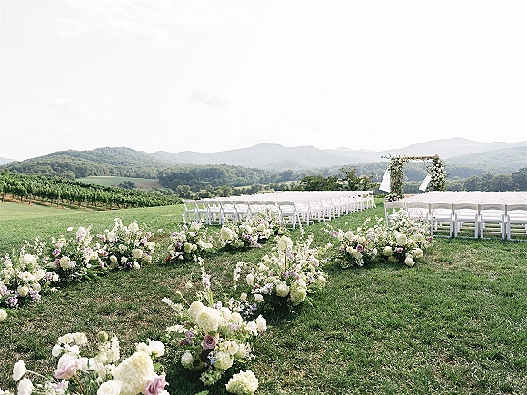 Ceremony setup for an outdoor wedding ceremony with white folding chairs, floral aisle arrangements, and a draped wooden arch on a vineyard lawn with mountains