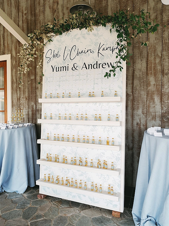 Wedding escort display with a calligraphy sign and greenery garland, shelves of place cards and mini bottles against a rustic wood wall