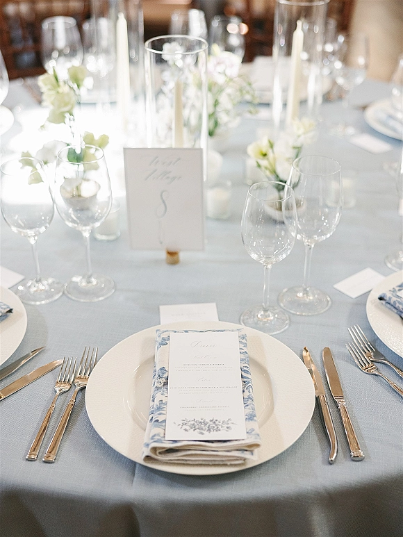 Reception tablescape with blue and white place settings on a light blue tablecloth, white flowers and candles in a softly lit room