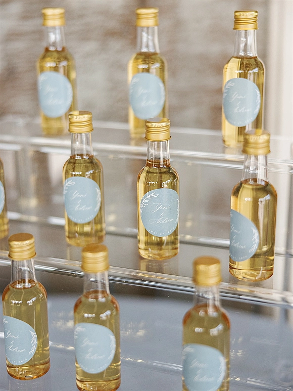 Wedding favor bottles on clear acrylic risers with gold screw caps and round labels arranged neatly on a tabletop against a neutral wall