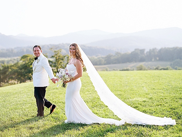 Couple portrait of bride and groom holding hands, bride with bouquet and long veil, walking through a mountain meadow backdrop