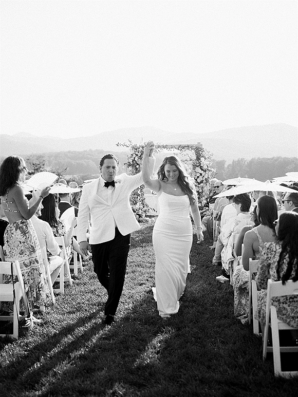 Wedding recessional as bride and groom hold hands with raised arms, her strapless gown and his white jacket, guests with parasols, mountain view backdrop