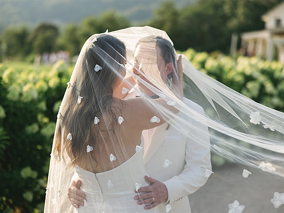 Wedding kiss portrait with veil over face kiss as bride and groom embrace under a floral appliqué veil on an outdoor walkway by hedges and mountains
