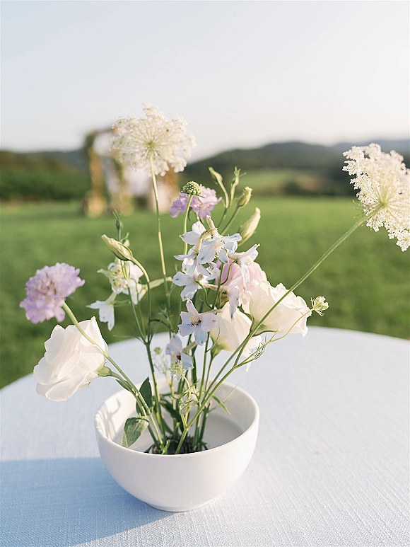 Wedding centerpiece of wildflowers in a white bowl vase on a tablecloth, with hills, sky, and a blurred ceremony arch behind