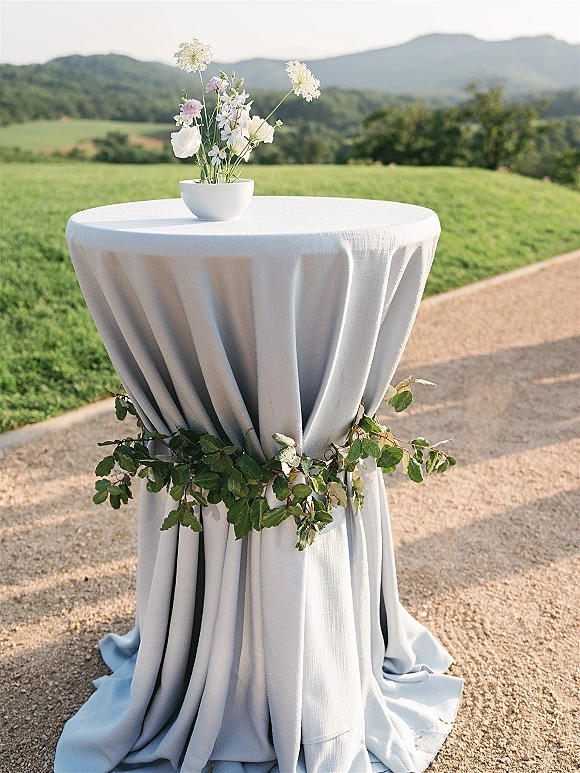 Cocktail table decor with a white tablecloth and small floral arrangement in a white bowl vase, set outdoors with mountains beyond trees