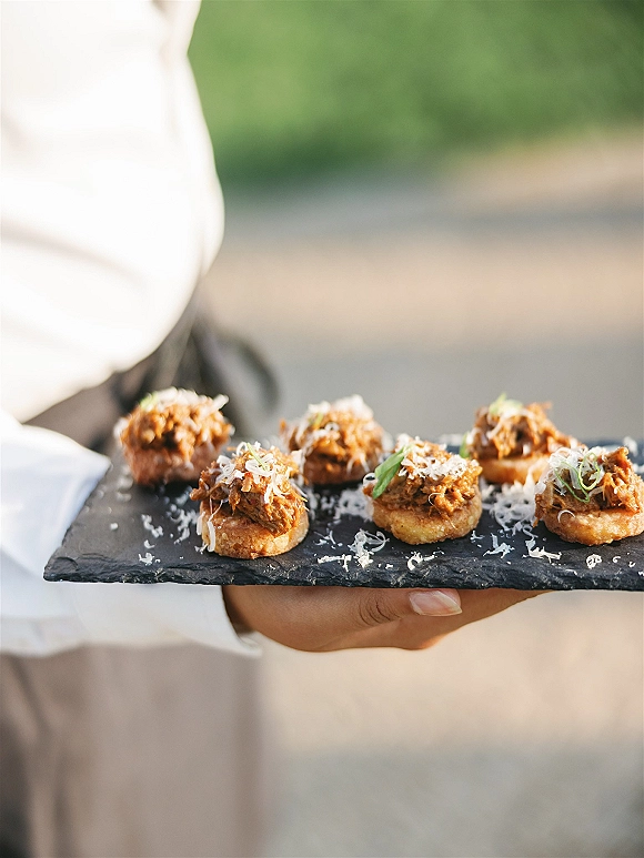 Wedding appetizers on a slate serving platter, bite-size canapés with shredded cheese and green onion garnish at an outdoor lawn cocktail hour