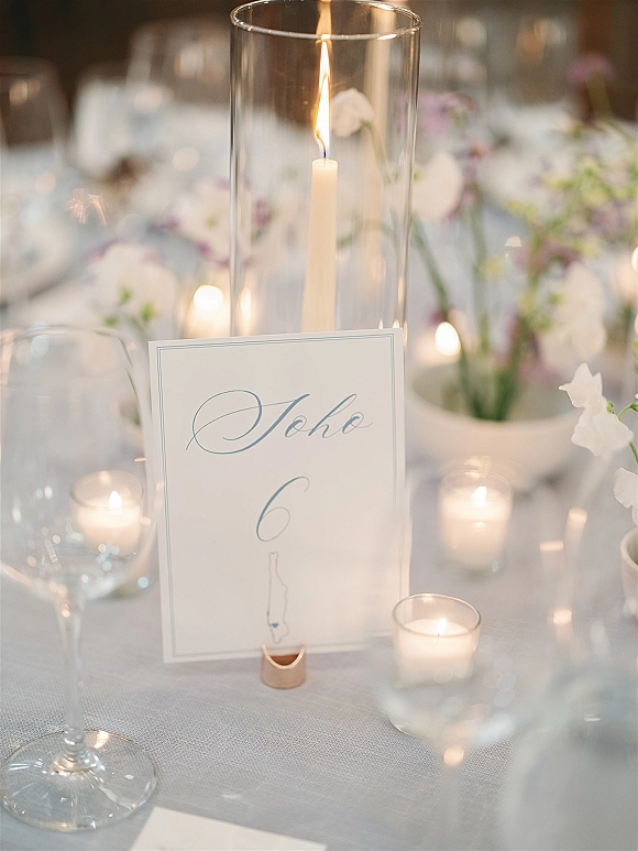 Wedding table number card with calligraphy in a gold holder, surrounded by taper and votive candles, wine glasses, and floral centerpiece