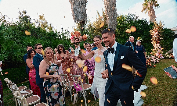 Wedding recessional as bride and groom walk the aisle through a rose petal toss, bouquet raised, under a floral arch in a palm-lined garden lawn