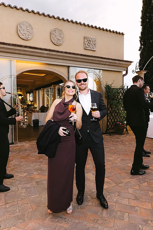 Wedding guest portrait of a stylish couple in sunglasses holding cocktails on an outdoor patio by a stucco building with arched doorway