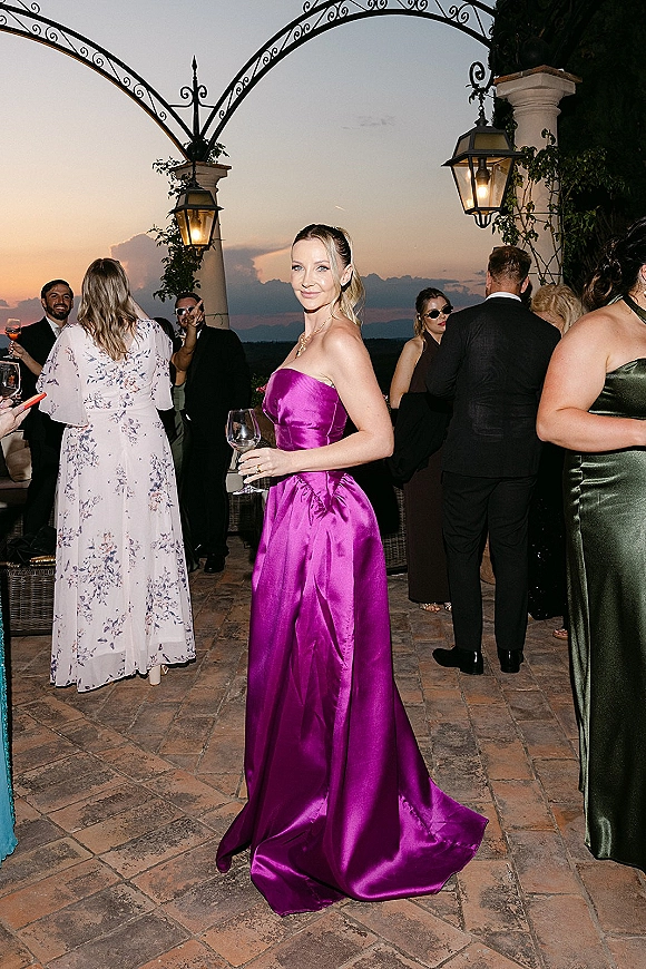Wedding guest outfit in a purple satin gown holding a wine glass, wearing gold jewelry, on a stone terrace at sunset under lanterns