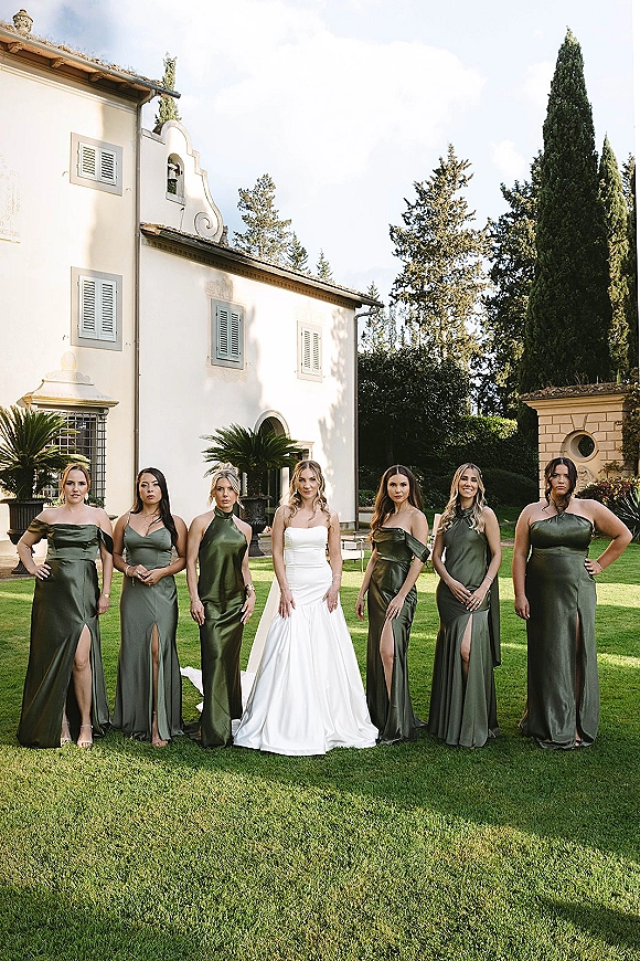 Bride with bridesmaids in olive green satin dresses posing on a lawn, bride in a strapless gown before a villa facade and trees