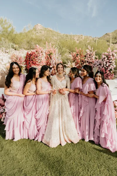 Bridesmaids group photo with bride and bridesmaids in pink dresses, laughing by a floral stage with desert mountains and cacti behind