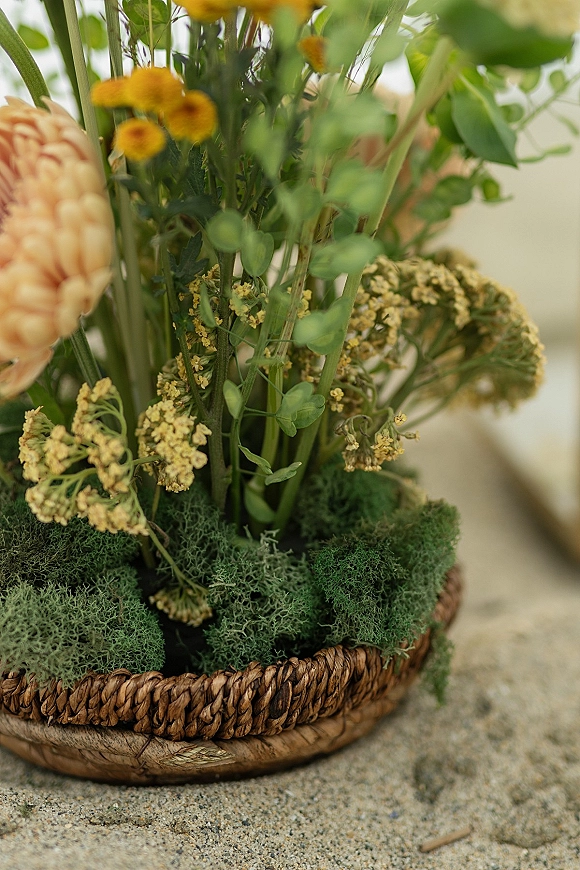 Wedding centerpiece with a floral centerpiece basket, moss and greenery topped with yellow wildflowers on sand against a neutral backdrop