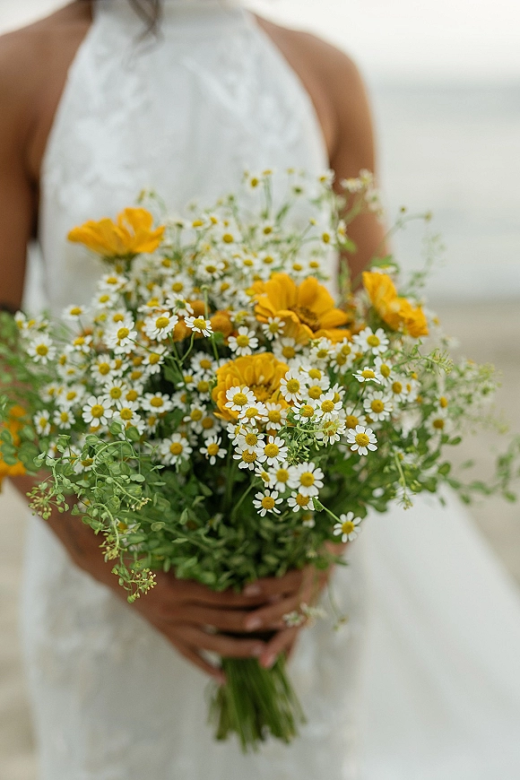 Bridal bouquet of chamomile daisies and yellow wildflowers with greenery held against a white halter wedding dress indoors by a neutral wall
