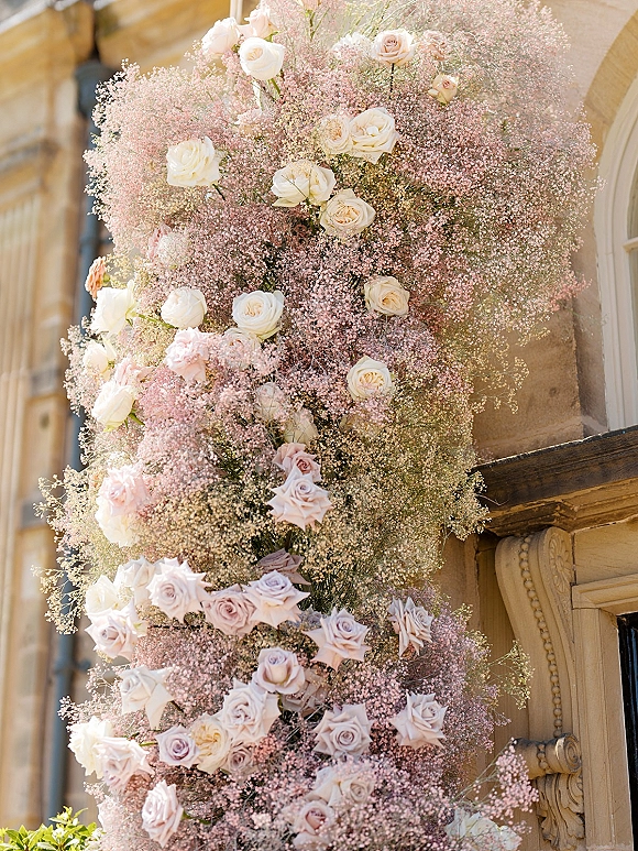 Wedding floral installation with roses and baby's breath cascading around a doorway on a stone building facade, lush greenery accents