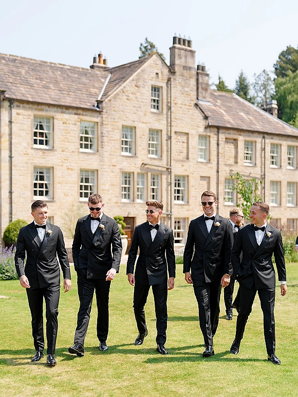 Groomsmen portrait of men in black tuxedos walking on a lawn, some in sunglasses, with a stone manor house and blue sky behind