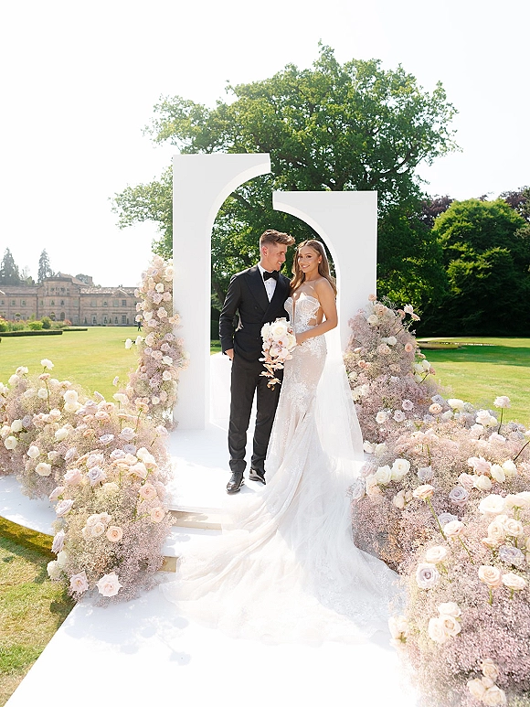 Couple portrait of bride holding a blush rose and baby's breath bouquet beside groom in black tuxedo under a modern white garden arch