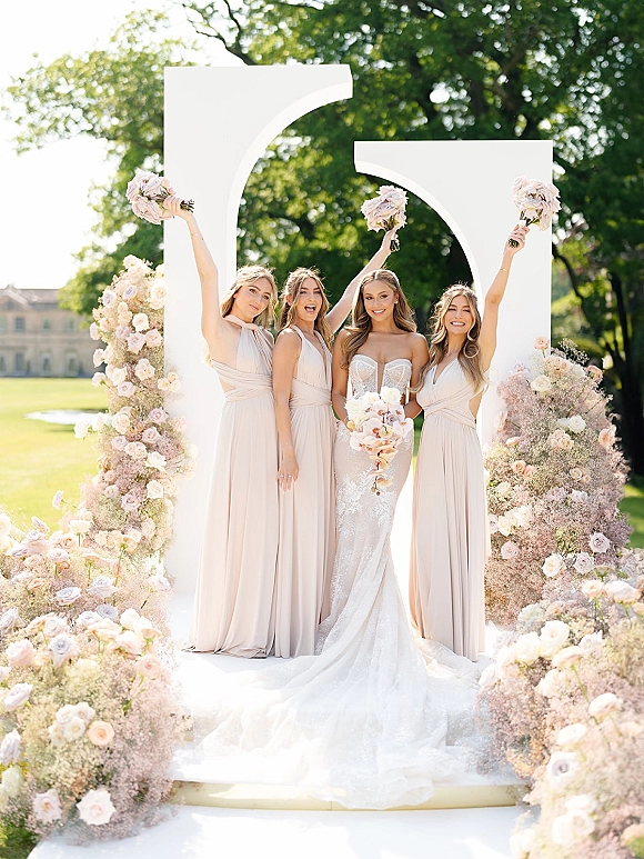 Bride with bridesmaids posing in blush dresses, raising bouquets beside a white floral arch on a sunny lawn with trees behind