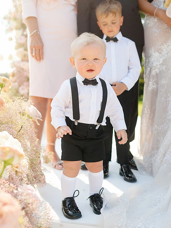 Ring bearer portrait of a toddler in black suspenders and bow tie walking with the wedding party along an outdoor greenery-lined walkway