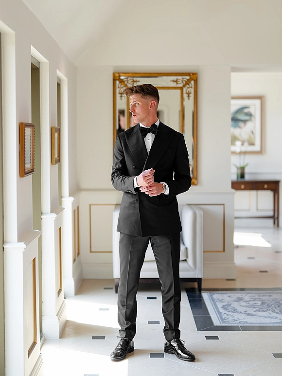 Groom portrait in a black tuxedo, bow tie and wristwatch, standing in a bright hallway with ornate mirror and framed artwork backdrop