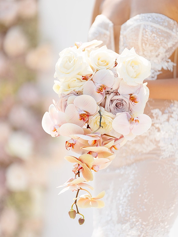Bridal bouquet with cascading orchid bouquet of white and blush roses, trailing orchids, held against a lace wedding dress in soft greenery bokeh