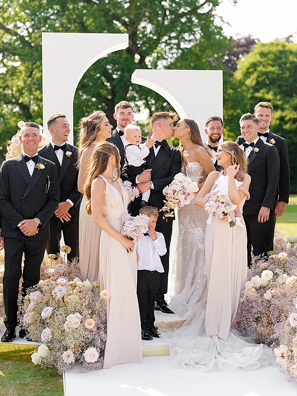 Wedding party portrait of bride and groom kissing with bridesmaids holding blush rose bouquets and groomsmen in tuxedos on a garden lawn