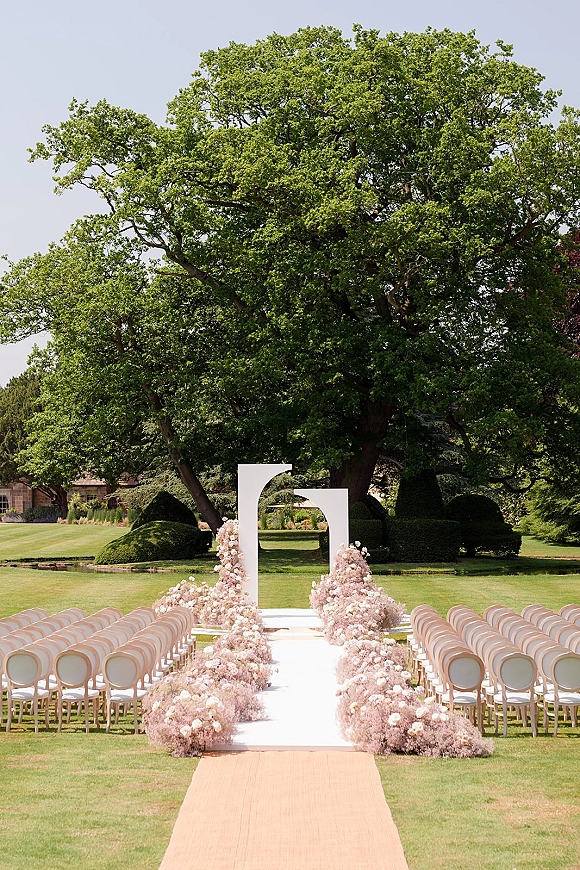 Ceremony setup with a white geometric arch, floral aisle arrangements, and a white runner on a lawn beneath a large tree