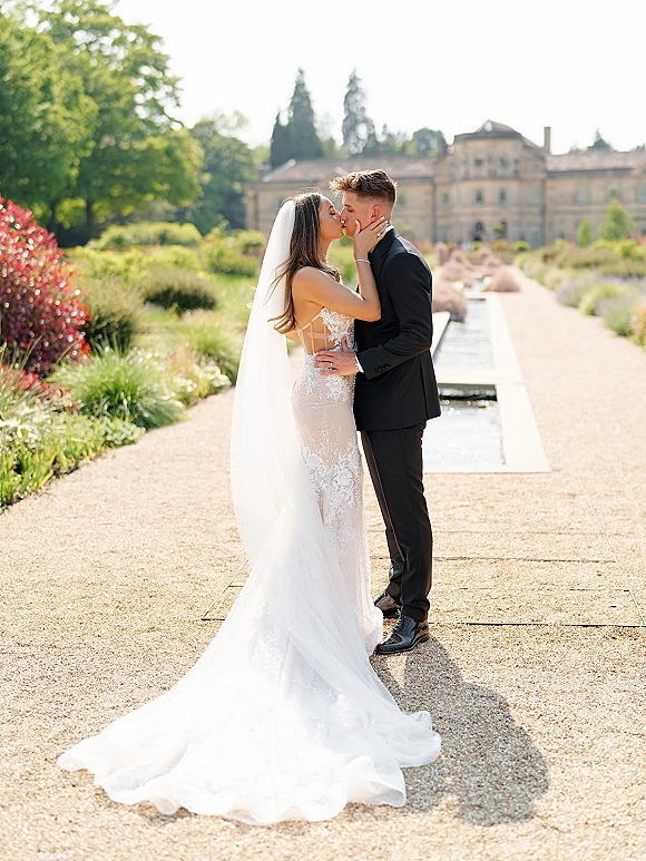 Wedding kiss portrait of bride and groom kissing, her veil blowing as she holds his face on a gravel path by a reflecting pool.