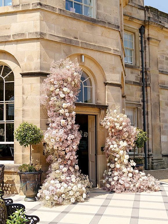 Wedding entrance florals framing floral entrance pillars with blush roses and baby’s breath at a stone manor doorway with topiary planters