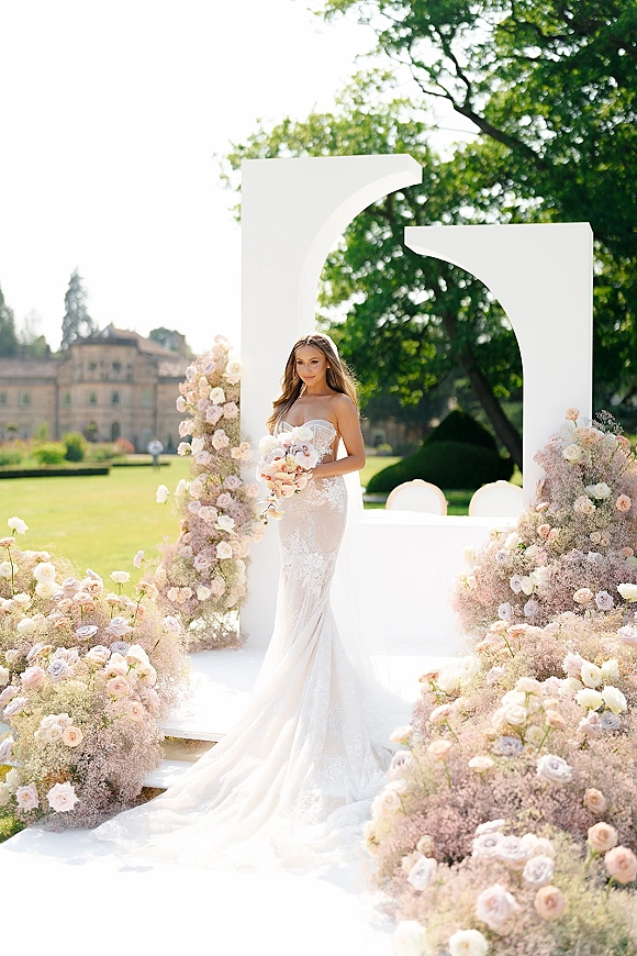 Bridal portrait of a bride in a strapless lace wedding dress with long train, holding a pastel bouquet by a white garden arch at an estate lawn ceremony