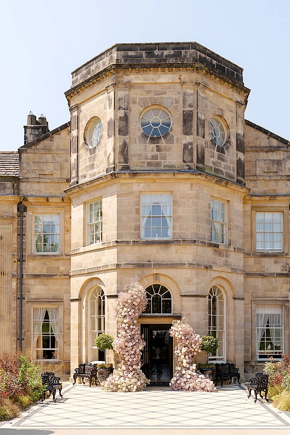 Wedding entrance decor with a floral entrance installation of pink and white blooms framing an arched stone manor doorway in a courtyard