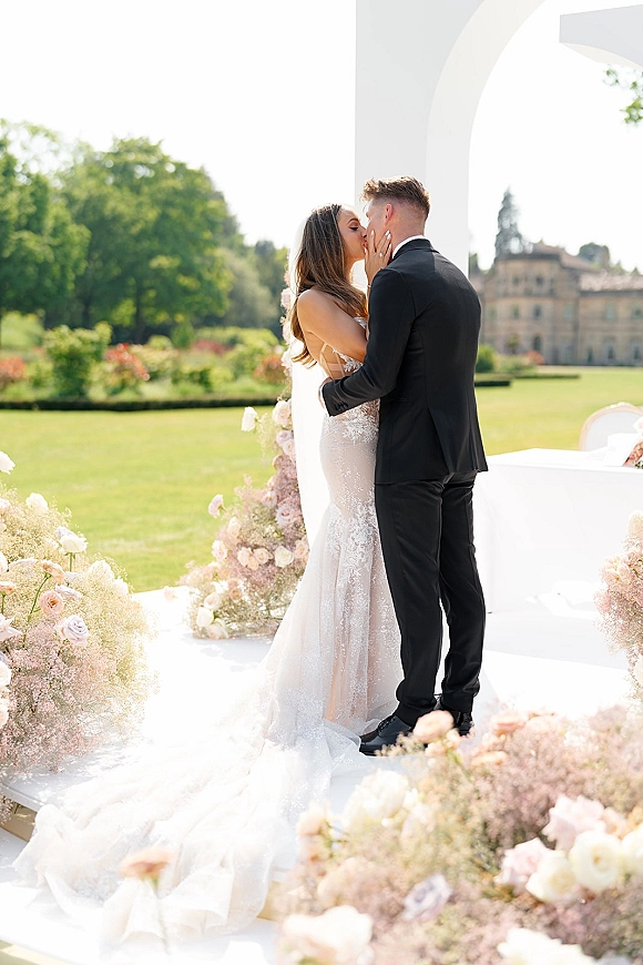 Wedding kiss as bride in lace dress and long veil kisses groom in black tuxedo beneath a floral arch on a garden lawn by the manor house