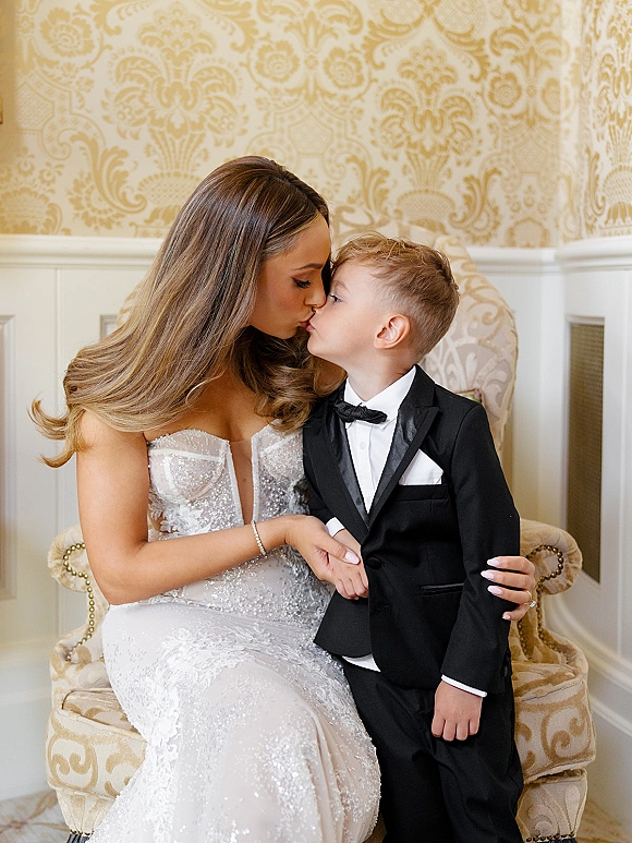 Wedding family portrait of a bride kissing child in a sparkly strapless gown beside a little boy in a tux, against damask wallpaper indoors