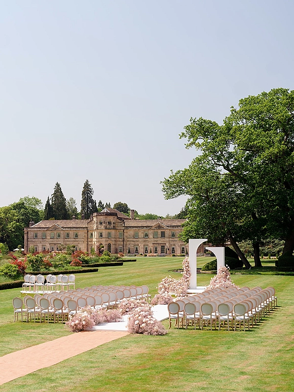 Ceremony setup for an outdoor wedding ceremony with a white arch, floral installations, and aisle runner on a manor house lawn