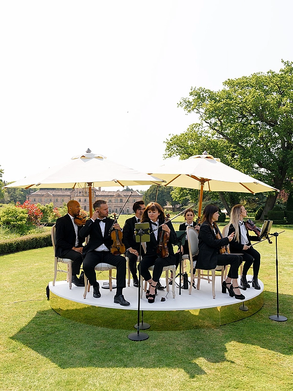 Wedding string quartet outdoor wedding musicians in formal black attire on a white stage with microphones, music stands, and umbrellas by a manor lawn