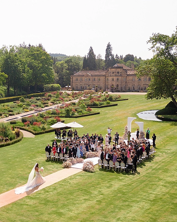 Outdoor wedding ceremony with a bride walking down the long aisle runner toward white arch columns in a formal estate garden setting