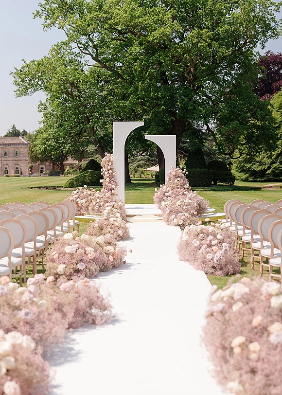 Ceremony aisle decor with an outdoor ceremony aisle, white aisle runner and blush floral ground arrangements leading to a curved arch on a manicured lawn