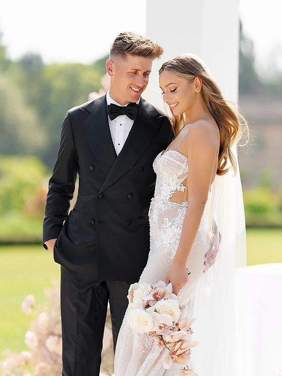 Couple portrait of bride in strapless lace dress and veil holding orchid bouquet beside groom in black tuxedo on a green lawn by a white column