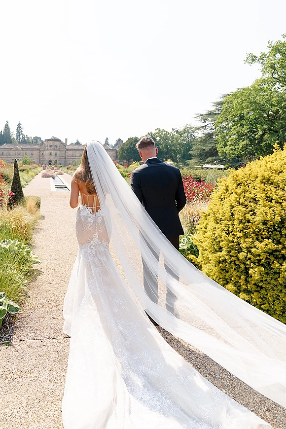 Couple portrait of bride and groom walking away, her cathedral-length wedding veil trailing on a gravel path in a formal garden estate