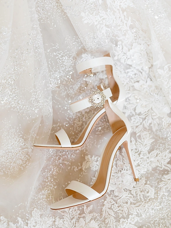 Bridal shoes, white wedding heels with crystal buckle and ankle straps resting on lace and sequin tulle wedding dress fabric flatlay