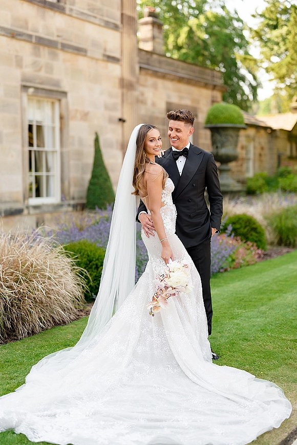 Couple portrait of bride and groom posing, groom hugging her as she looks over shoulder, long veil and bouquet by stone manor garden lawn