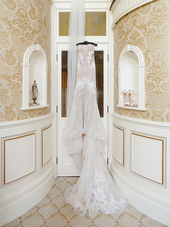 Wedding dress with lace wedding dress details hangs on a hanger, with veil and nude strappy heels against ornate wallpaper and doors