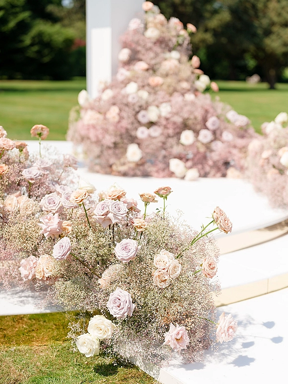 Ceremony aisle flowers in grounded clusters of pastel roses and baby’s breath lining a white platform on a green garden lawn with trees