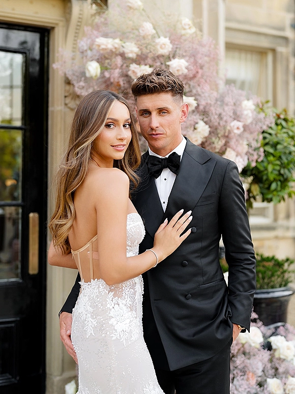 Couple portrait of bride in a strapless lace wedding dress with bracelet and groom in black tuxedo before a floral backdrop on stone facade