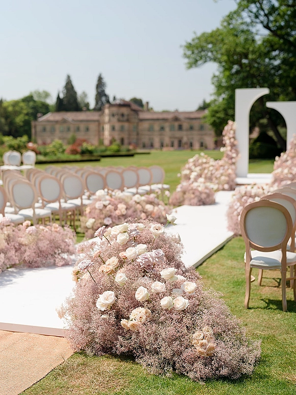Ceremony aisle decor with a white aisle runner lined with blush and white rose ground florals leading to a curved arch on a manor lawn under blue sky