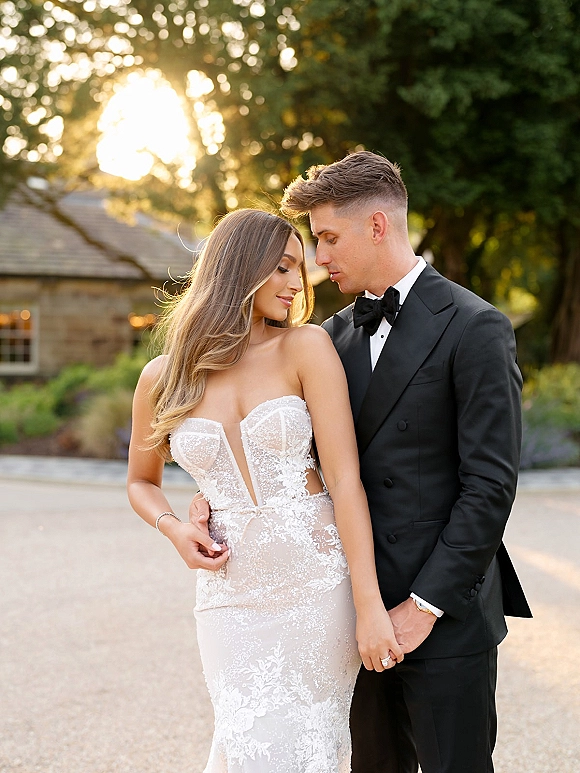 Couple portrait of bride and groom embrace, her strapless lace dress and his black bow tie framed by garden trees and sun flare