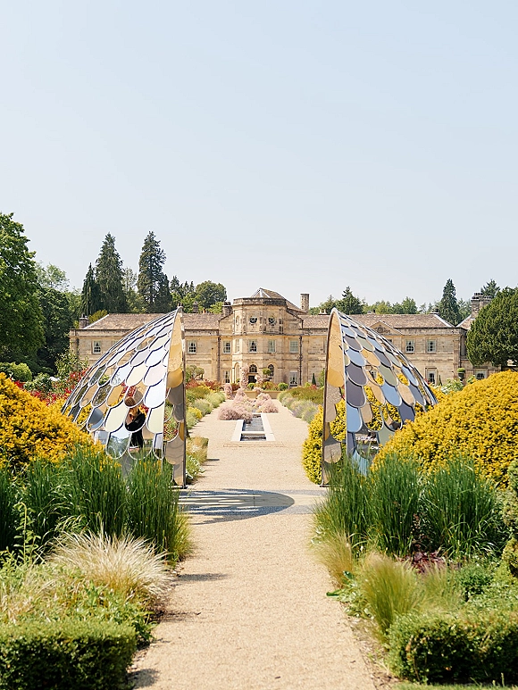 Garden wedding venue with a gravel pathway leading past modern metal pavilions and hedges to a historic manor house under blue sky