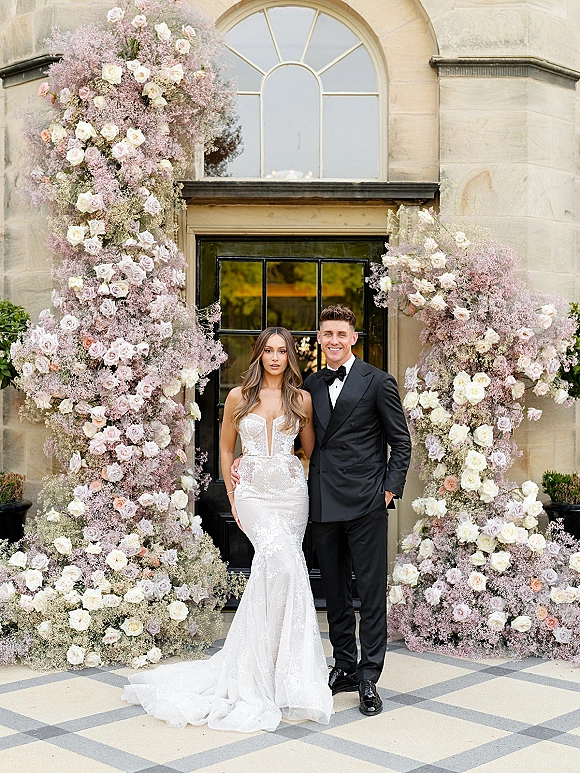Couple portrait of bride in strapless lace dress and groom in black tuxedo under a rose and baby's breath arch at a stone entryway