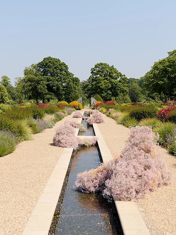 Garden walkway with a formal garden path beside a water rill, lined with lavender, ornamental grasses, and flowering shrubs under blue sky