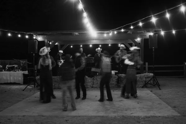 Wedding dance floor with live band wedding reception under string lights in a wooden pavilion at night, with hay bales and cowboy hats